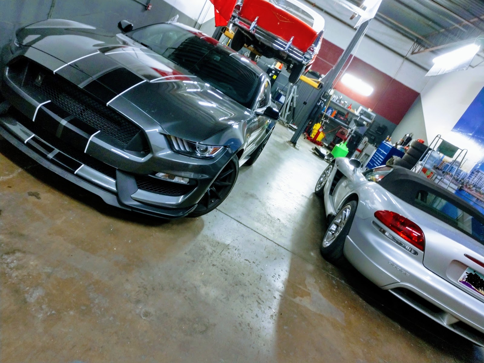 Inside the shop: a gray Mustang with racing stripes and a silver Viper convertible parked side by side while a red classic car sits high on the lift