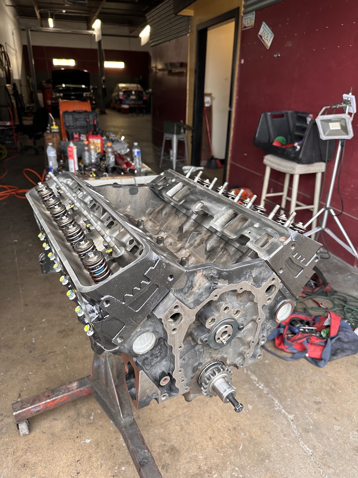 A V8 engine block on a rolling engine stand with valve springs exposed, in front of the shop's burgundy wall