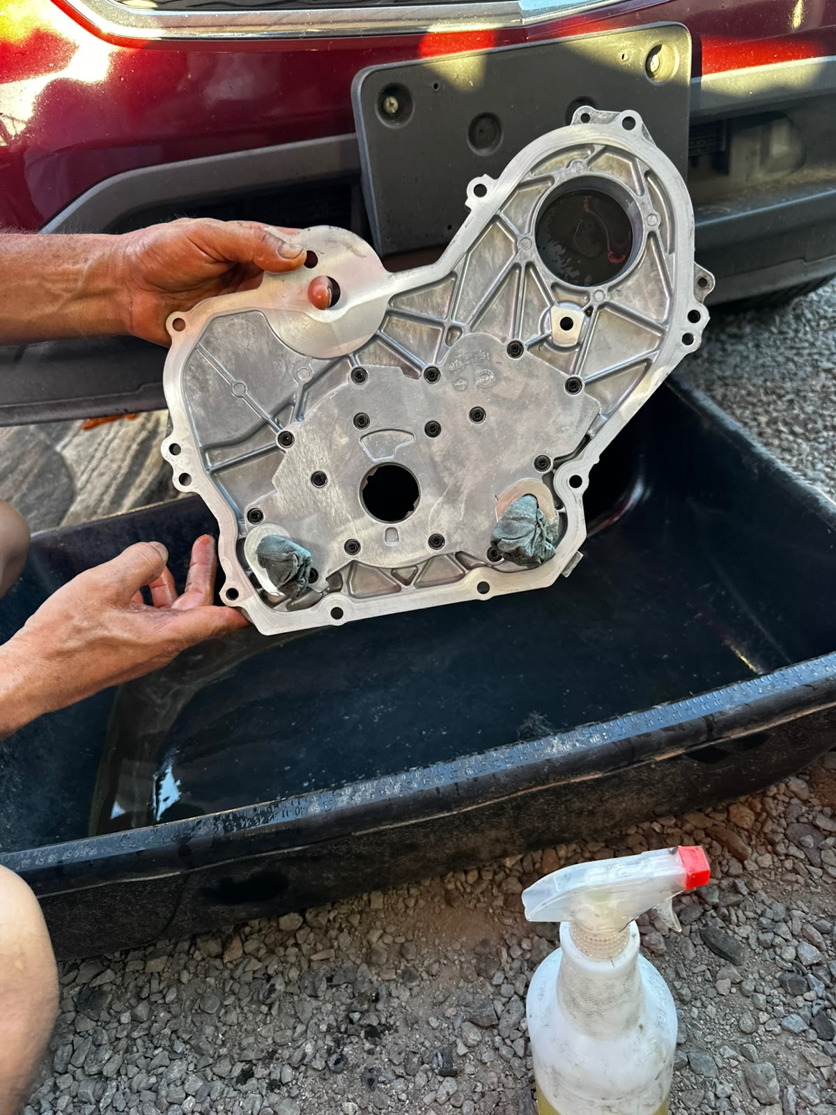 Hands holding a freshly degreased timing cover over a drain pan outdoors